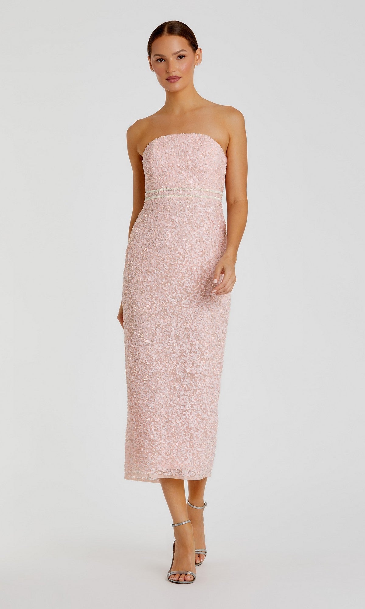 A woman poses in the Tea-Length Party Dress: Mac Duggal 94200—a blush, sequined midi design featuring a strapless neckline, fitted silhouette, and subtle pink pearl-embellished details. She completes the elegant look with silver high-heeled sandals against a plain light background.